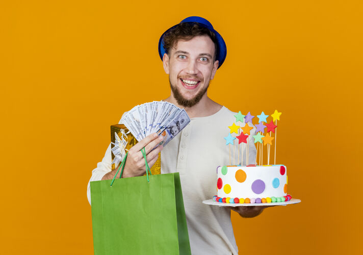 盒子快乐的年轻帅哥斯拉夫党家伙戴着党帽子拿着礼品盒钱纸袋和生日蛋糕与明星看着相机隔离在橙色背景与复制空间礼物生日小伙子