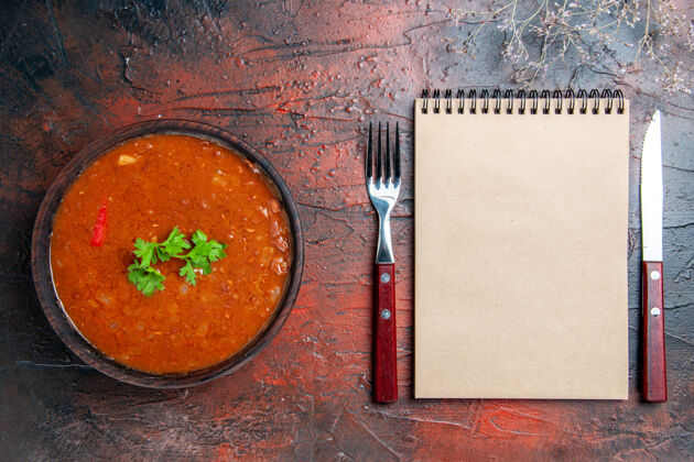 碗经典的西红柿汤在一个棕色的碗和勺子与叉子和刀子和笔记本在混合颜色的桌子上晚餐打火机汤