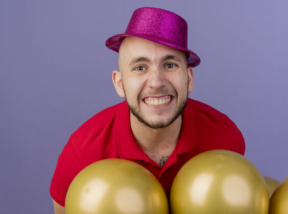 小伙子微笑的年轻英俊的斯拉夫党人戴着党帽站在气球后面看着紫色背景上孤立的相机特写镜头风景特写派对