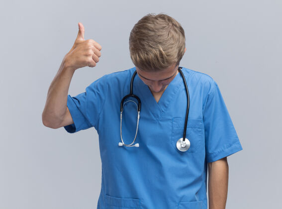 男性低着头的年轻男医生穿着医生制服 听诊器在白色的墙上显示拇指向上隔离展示拇指年轻