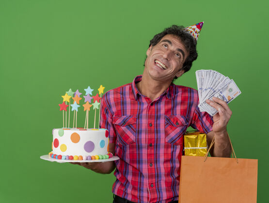 蛋糕快乐的中年白种人聚会男子戴着生日帽拿着生日蛋糕纸袋礼包和钱孤立地仰望绿色背景纸聚会男人