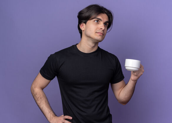 小伙子想着年轻帅哥穿着黑色t恤拿着一杯咖啡把手放在屁股上隔离在紫色的墙上年轻杯子臀部