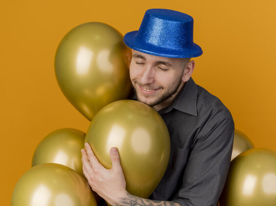 抓住微笑着的年轻英俊的斯拉夫党人戴着党的帽子站在气球抓住一个闭着眼睛孤立在橙色背景站着小伙子派对