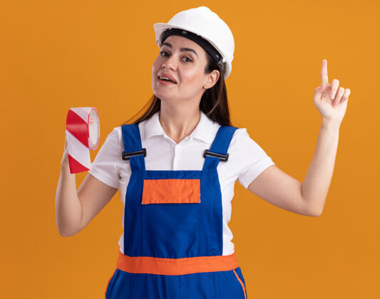 女人令人印象深刻的年轻建筑妇女在统一举行管道胶带点在橙色墙上孤立制服管道磁带