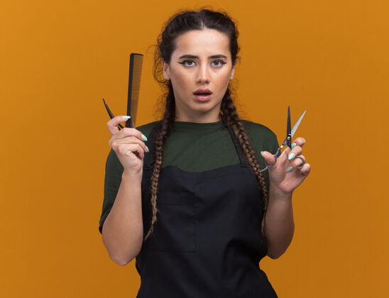 理发师印象深刻的年轻女理发师在制服举行理发工具孤立的橙色墙上与复制空间印象手持女