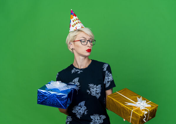 礼物可疑的年轻金发派对女孩戴着眼镜和生日帽拿着礼品盒看着其中一个孤立的绿色背景生日拿着金发