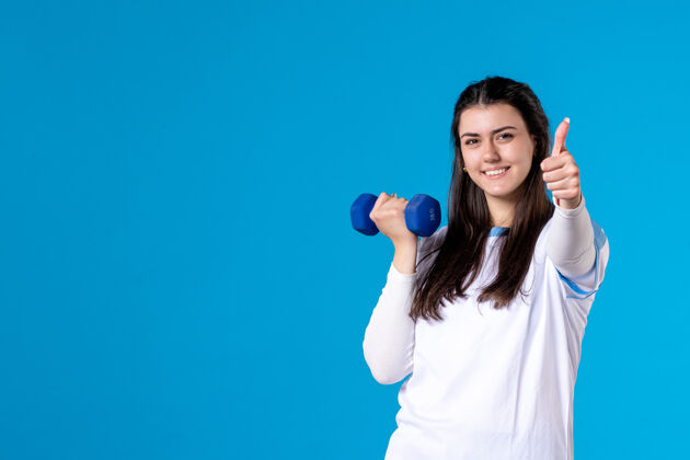 身体正面图：年轻女性在蓝色墙上用蓝色哑铃锻炼人微笑前面