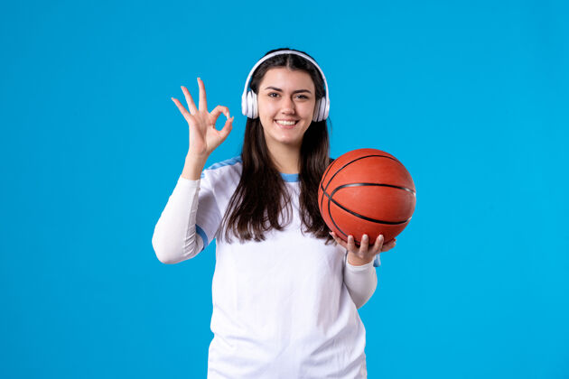 成人正面图：戴着耳机拿着篮球的年轻女性站在蓝色的墙上年轻女性比赛年轻人