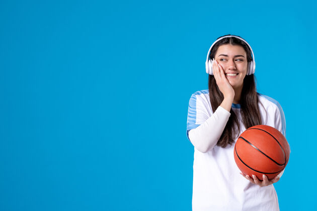 观点正面图：戴着耳机拿着篮球的年轻女性站在蓝色的墙上人成人比赛