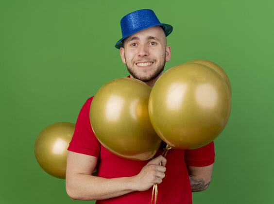 绿色面带微笑的年轻英俊的斯拉夫党人戴着党的帽子拿着气球看着隔离在绿色背景上的相机微笑斯拉夫人小伙子