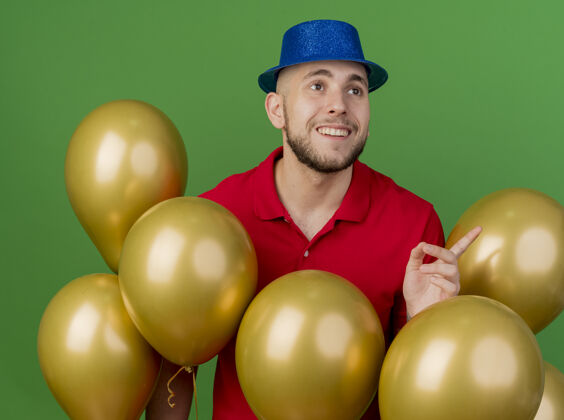 站着面带微笑的年轻英俊的斯拉夫党人戴着党的帽子站在气球后面看着并指着绿色背景上孤立的一面年轻人小伙子绿色