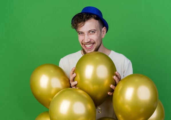 斯拉夫人快乐的年轻英俊的斯拉夫党人戴着党的帽子站在气球后面看着相机拿着绿色背景上孤立的气球小伙子欢乐抱着