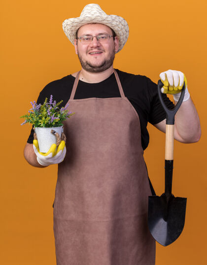 园艺微笑着的年轻男园丁戴着园艺帽和手套拿着铁锹 花盆里的花被隔离在橙色的墙上男黑桃花园