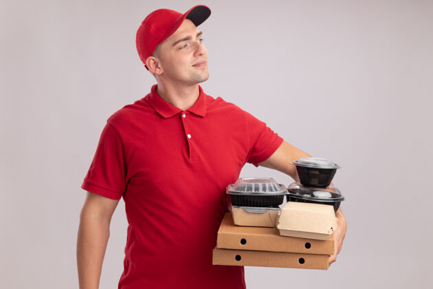 送货高兴地看着旁边的年轻送货员穿着制服 戴着帽子 把食品容器放在隔离在白墙上的比萨饼盒上男人壁板制服