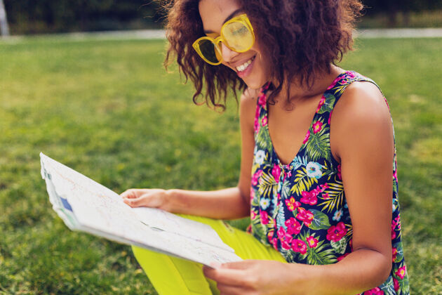太阳镜年轻时尚的黑人女子在公园享受夏日时尚的乐趣 五颜六色的时髦装扮 坐在草地上 旅行者拿着地图积极肖像酷