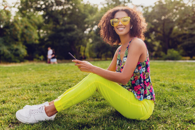 混血儿年轻时尚微笑的黑人女子用智能手机听音乐无线耳机在公园里玩得开心 夏日时尚多彩的风格 坐在草地上 戴着黄色太阳镜兴奋酷欢呼