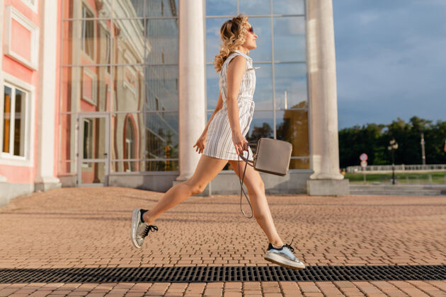 女性年轻迷人的时尚女子穿着运动鞋在城市街道上奔跑跳跃有趣夏天时尚风格的白色连衣裙戴着太阳镜和手提包优雅连衣裙姿势