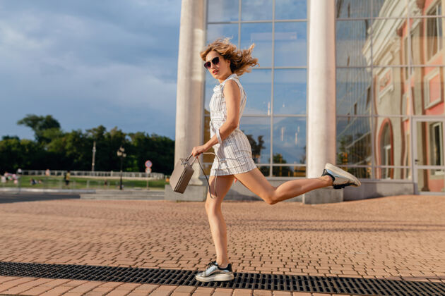 女孩年轻迷人的时尚女子穿着运动鞋在城市街道上奔跑跳跃有趣夏天时尚风格的白色连衣裙戴着太阳镜和手提包别致优雅城市
