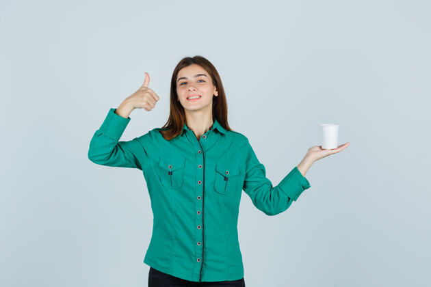 前面穿着衬衫的年轻女士拿着一杯塑料咖啡 同时竖起大拇指 看起来很愉快 正前方视图朋友时尚年轻