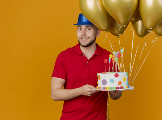 气球面带微笑的年轻帅哥斯拉夫党人戴着党的帽子拿着气球和生日蛋糕看着相机孤立的橙色背景与复制空间太空派对蛋糕