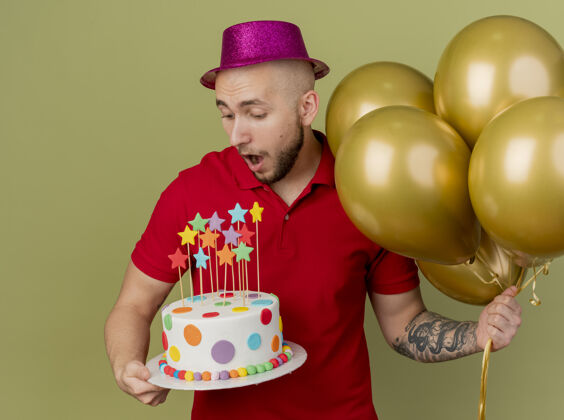 年轻人令人印象深刻的年轻英俊的斯拉夫党家伙戴着党的帽子拿着气球和生日蛋糕看着蛋糕孤立在橄榄绿的背景抱着派对印象