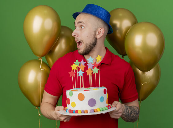 斯拉夫人印象深刻的年轻英俊的斯拉夫党人戴着党的帽子站在气球前拿着生日蛋糕看着绿色背景上孤立的一面小伙子派对前面