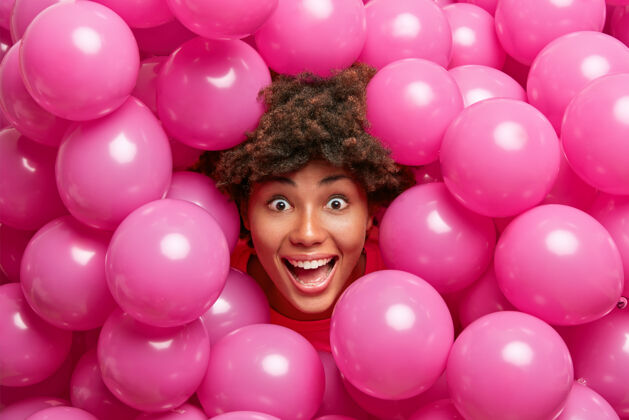 疯狂积极疯狂的黑皮肤女人看起来很高兴 得到惊喜 在节日里有乐趣的小充气玫瑰气球包围乐趣人类满意