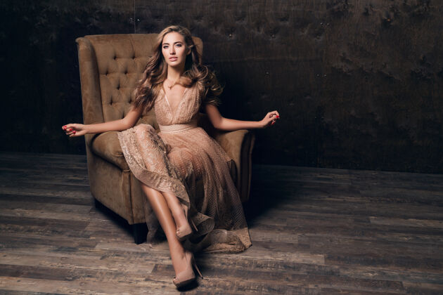 皮肤穿着闪亮蕾丝晚礼服的美丽性感模特坐在米色的椅子上衣服夜晚头发