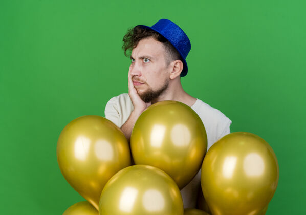 站着体贴的年轻英俊的斯拉夫党人戴着党的帽子站在气球后面 手放在脸上看着绿色背景上孤立的一面斯拉夫人手绿色