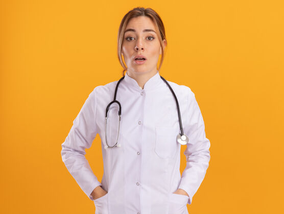 惊喜惊讶的年轻女医生穿着医用长袍 听诊器把手放在黄色墙上的口袋里听诊器女穿