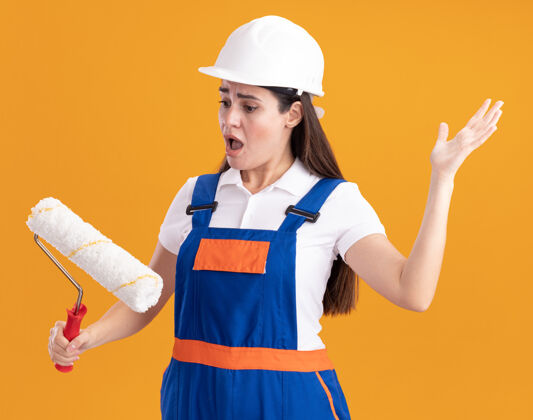 害怕一个穿着制服的年轻的建筑工人手里拿着刷子 看着隔离在橙色墙上的滚轴制服建筑工人刷子