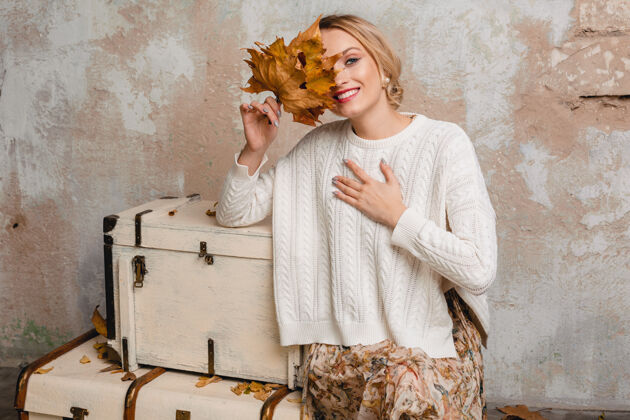 秋天迷人的微笑 穿着白色针织毛衣的时尚金发女人坐在手提箱上的肖像在街上的老式墙上肖像城市秋天