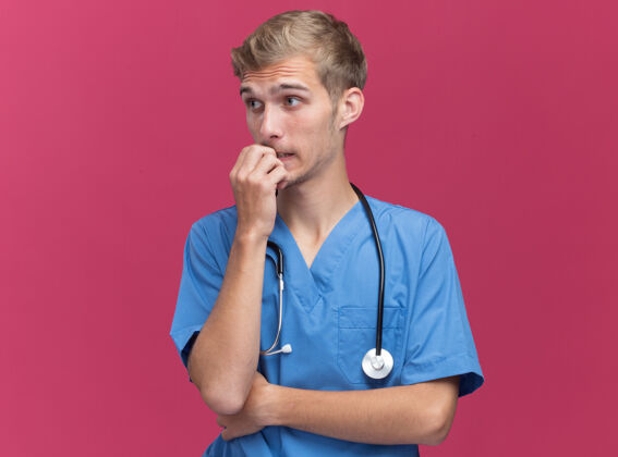 医生关心地看着身边年轻的男医生穿着医生制服用听诊器咬指甲隔离在粉红色的墙上年轻壁板咬
