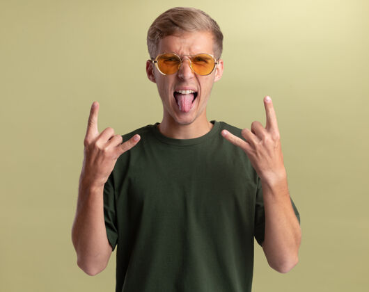 年轻年轻帅哥穿着绿色衬衫 戴着眼镜 露出舌头 在橄榄绿的墙上做山羊手势手势衬衫橄榄