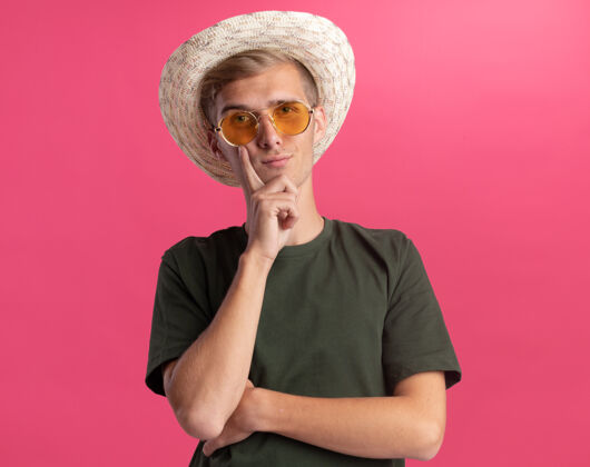 脸颊自信的年轻帅哥 穿着绿色衬衫 戴着眼镜 戴着帽子 手指放在脸颊上 隔离在粉红色的墙上年轻人手指自信