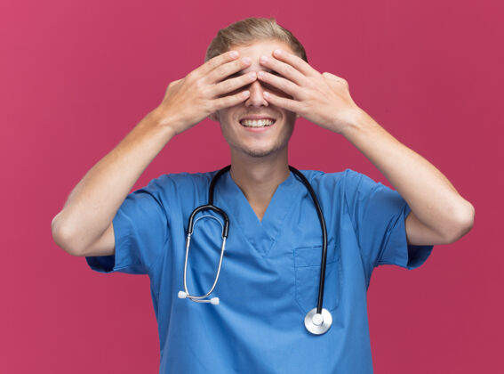 掩护面带微笑的年轻男医生穿着医生制服 听诊器盖着脸 手被隔离在粉红色的墙上医生年轻手