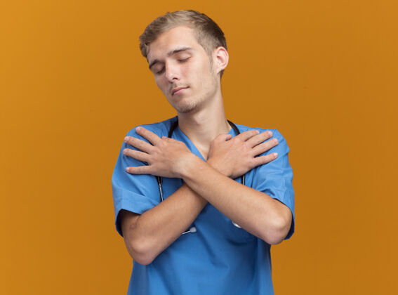 手年轻的男医生穿着医生制服 戴着听诊器 双手交叉放在隔离在橙色墙上的肩膀上制服听诊器放松