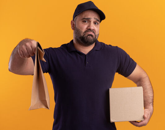 送货悲伤的中年送货员穿着制服 戴着帽子 拿着盒子 拿出纸食品包装隔离在黄色的墙上盒子男人中年