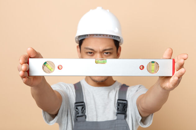 年轻严格的年轻建筑工人戴着安全帽和制服向摄像机伸出水平尺安全朝奶油