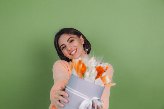 棉花花年轻女子穿着休闲桃色毛衣隔离在绿橄榄墙上手持橙白色花盒组成的棉花 吉普赛拉小麦和拉古鲁斯作为礼物开心惊喜年轻积极蝴蝶结