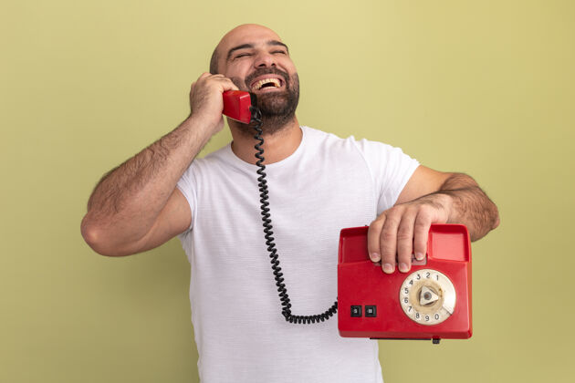 笑快乐的胡子男人穿着白色t恤拿着旧电话站在绿色的墙上大笑男人电话胡子