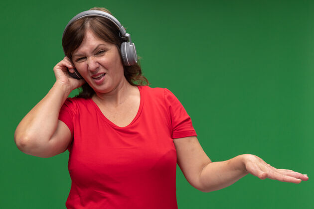 女人身穿红色t恤 头戴耳机 表情厌恶的中年妇女举起手臂站在绿色墙壁上提高立场厌恶