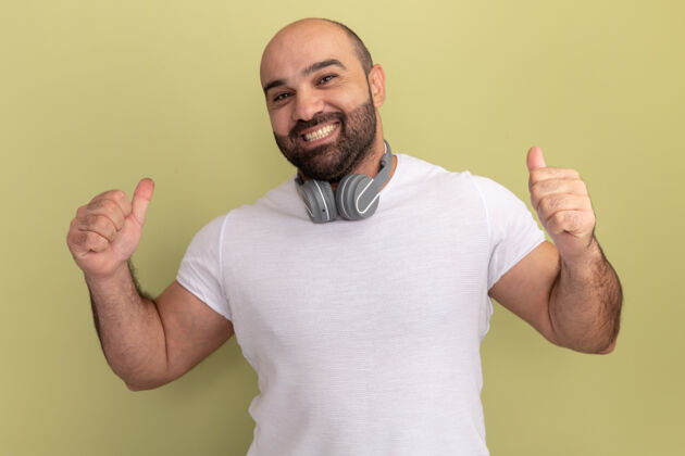 耳机一个留着胡子的男人 穿着白色t恤 戴着耳机 快乐而愉快地微笑着 竖起大拇指站在绿色的墙上胡子大拇指伙计