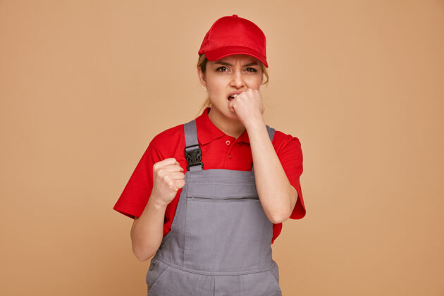 棕色焦急的年轻女建筑工人戴着帽子和制服握紧拳头咬另一个拳头握紧施工焦虑