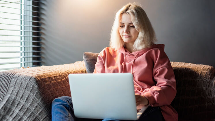 房子年轻的内容创造者金发女孩坐在靠近窗户的沙发上 坐在笔记本电脑上在家工作电子邮件彩色剪辑