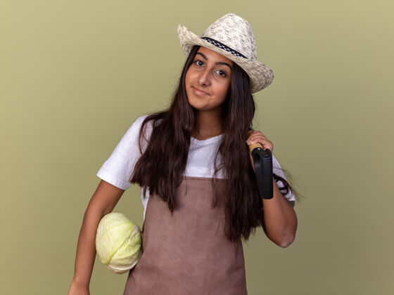年轻穿着围裙 戴着夏帽的年轻园丁女孩 手里拿着卷心菜和铲子 脸上带着微笑站在绿色的墙上铲子卷心菜花园
