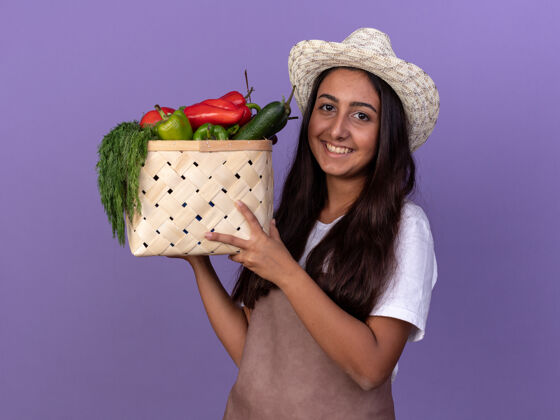 年轻穿着围裙 戴着夏帽 拿着装满蔬菜的箱子的年轻园丁女孩站在紫色的墙上 脸上洋溢着幸福的笑容充分欢呼围裙