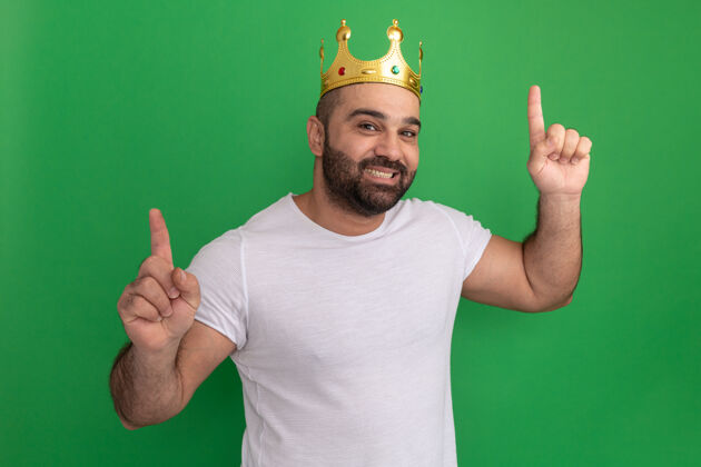 目录满脸胡须 身穿白色t恤 头戴金色皇冠的男子 快乐而积极地用食指指着绿色的墙壁男人手指皇冠