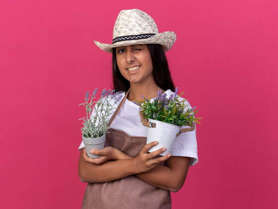 花盆快乐的年轻园丁女孩围着围裙 戴着夏帽 手里拿着盆栽植物 站在粉红色的墙上微笑着眨眼花园年轻人看台