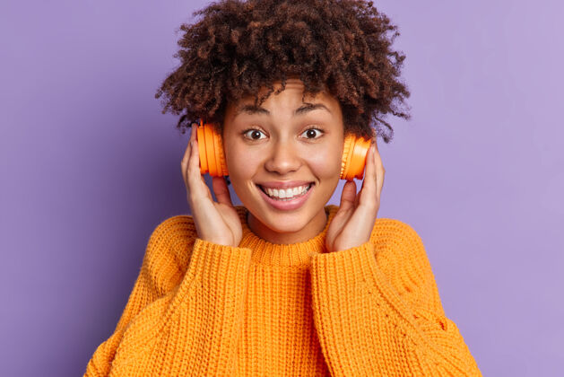 立体声美丽快乐的非洲裔美国卷发女孩的头像手放在耳机上微笑着牙齿穿针织毛衣的姿势高兴无忧无虑音乐
